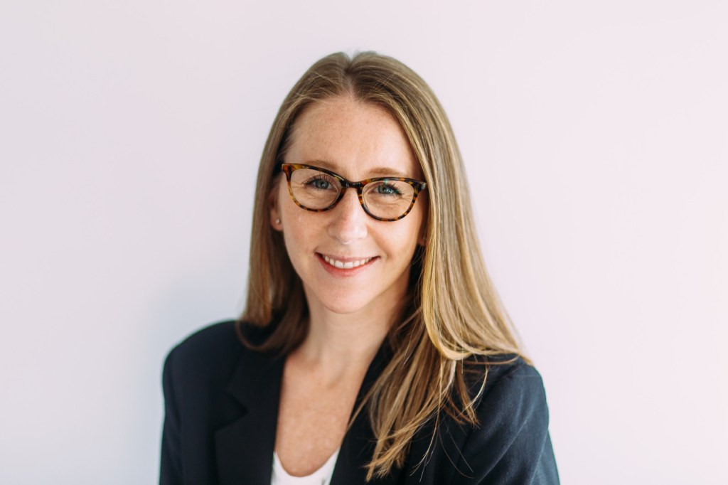 Cait Porte, smiling, in tortoiseshell glasses and a navy blazer